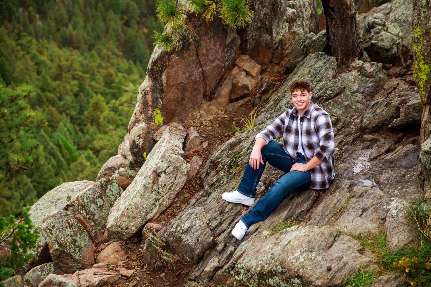 A person in plaid shirt and jeans sits casually on a rocky mountain ledge surrounded by evergreen forest.