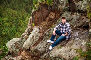 A person in plaid shirt and jeans sits casually on a rocky mountain ledge surrounded by evergreen forest.