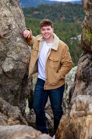 A person in a brown jacket and white shirt poses among rocky outcrops with forested mountains in the background.