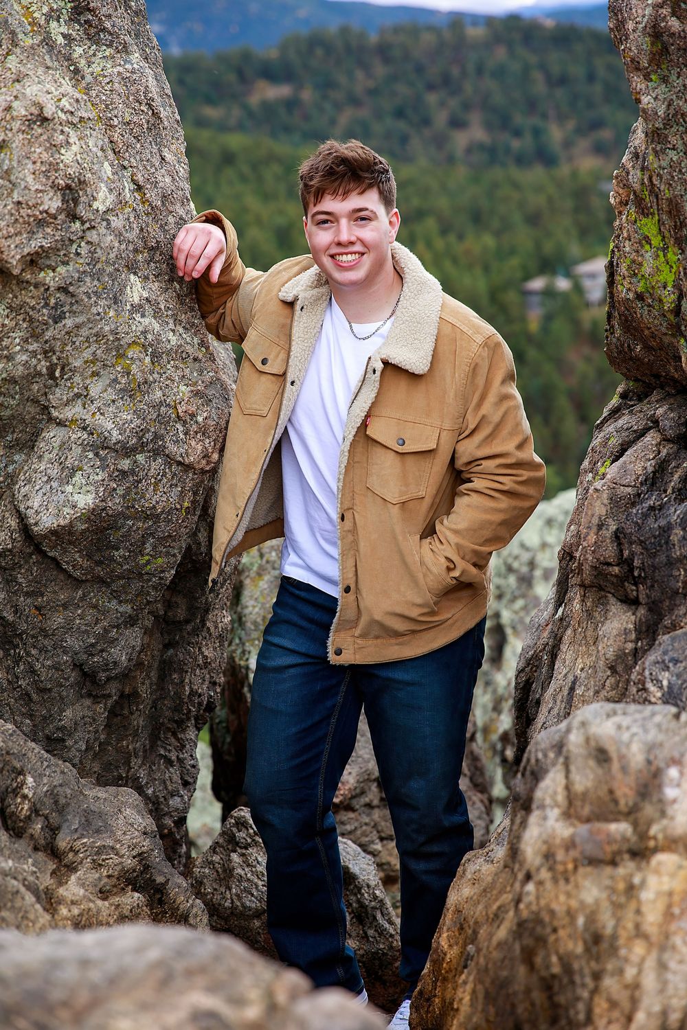 A person in a brown jacket and white shirt poses among rocky outcrops with forested mountains in the background.
