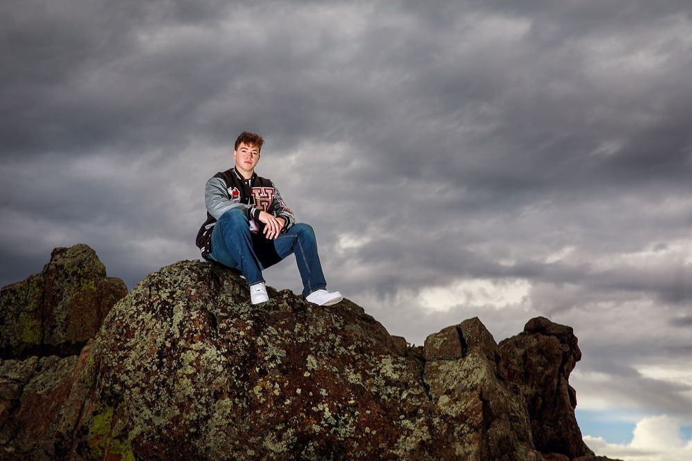 A person sits contemplatively on a rocky outcrop against dramatic storm clouds in the sky.