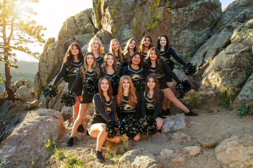 Cheerleaders in black uniforms pose together on rocky mountain terrain at sunset.