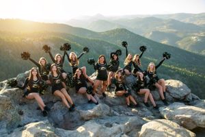 Group of cheerleaders raising pom poms while sitting on mountain rocks with scenic vista behind.