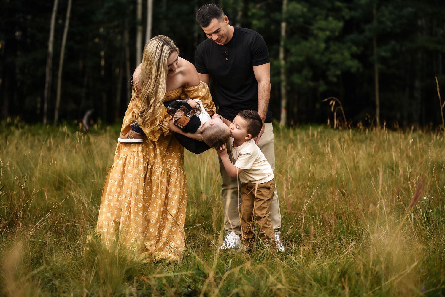 Happy family with toddler son playing in golden meadow with forest background during outdoor session.