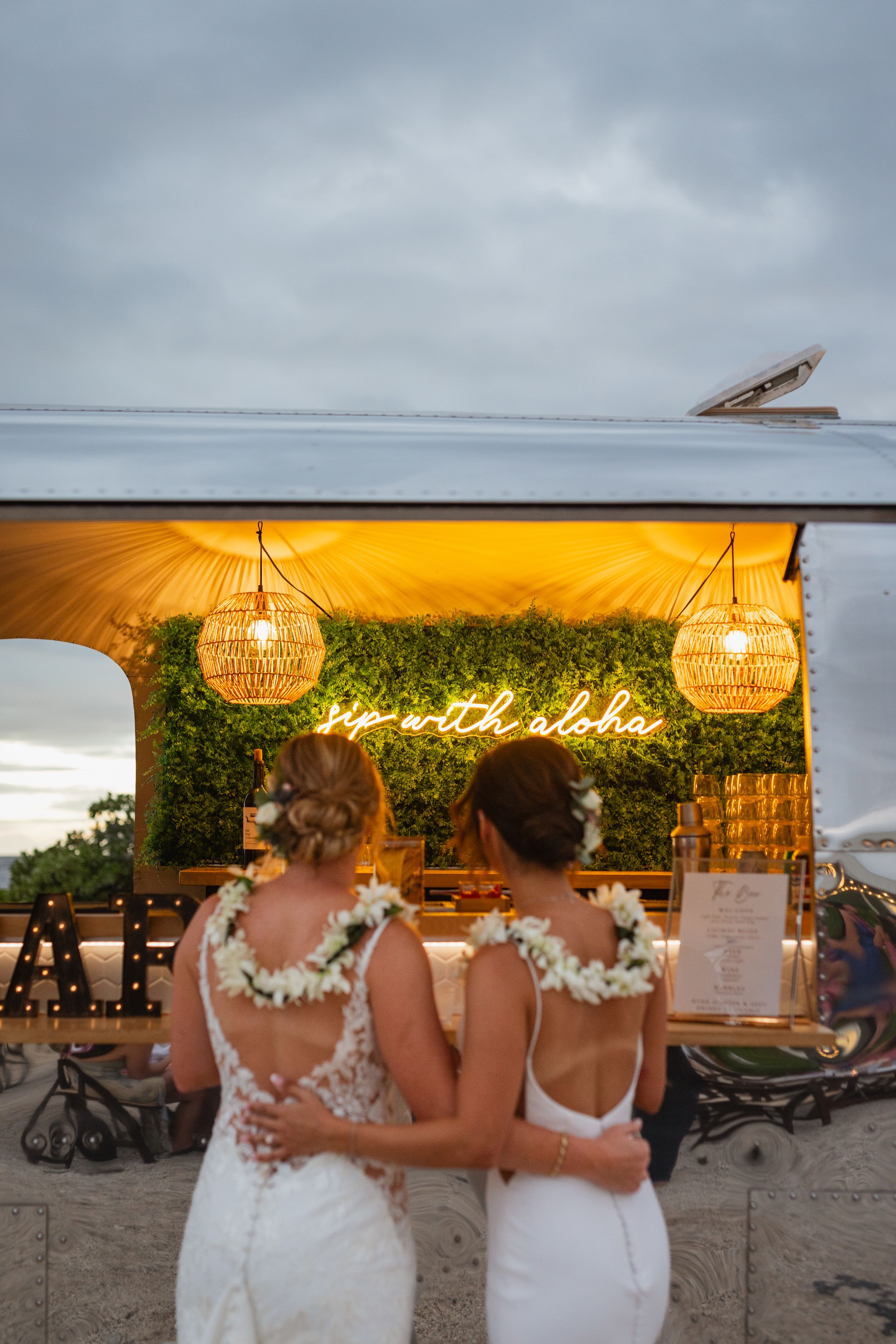 Two brides embrace while standing before a glowing bar truck with a sip with aloha neon sign.