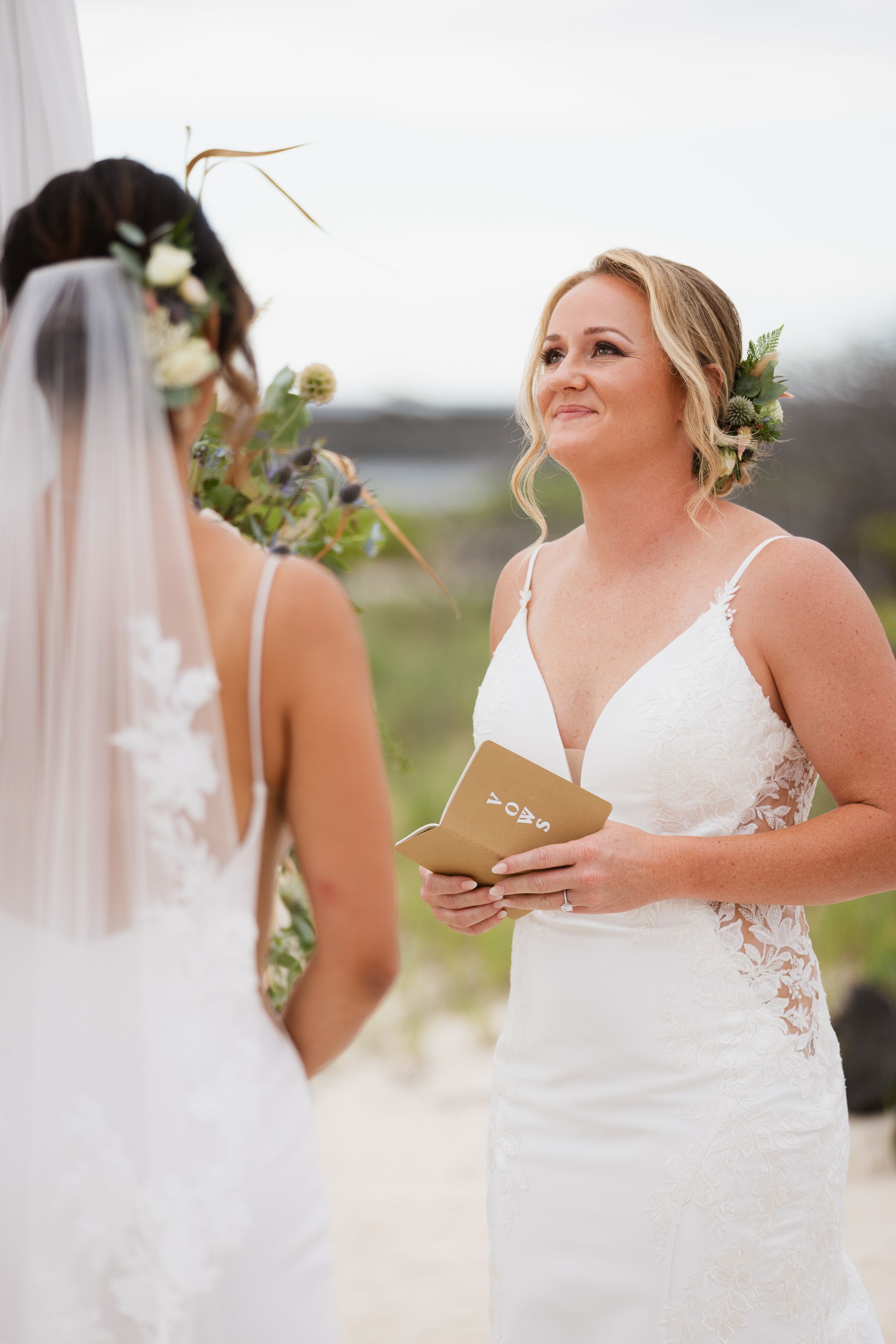 Blonde bride reads wedding vows while facing her partner during an outdoor beach ceremony.