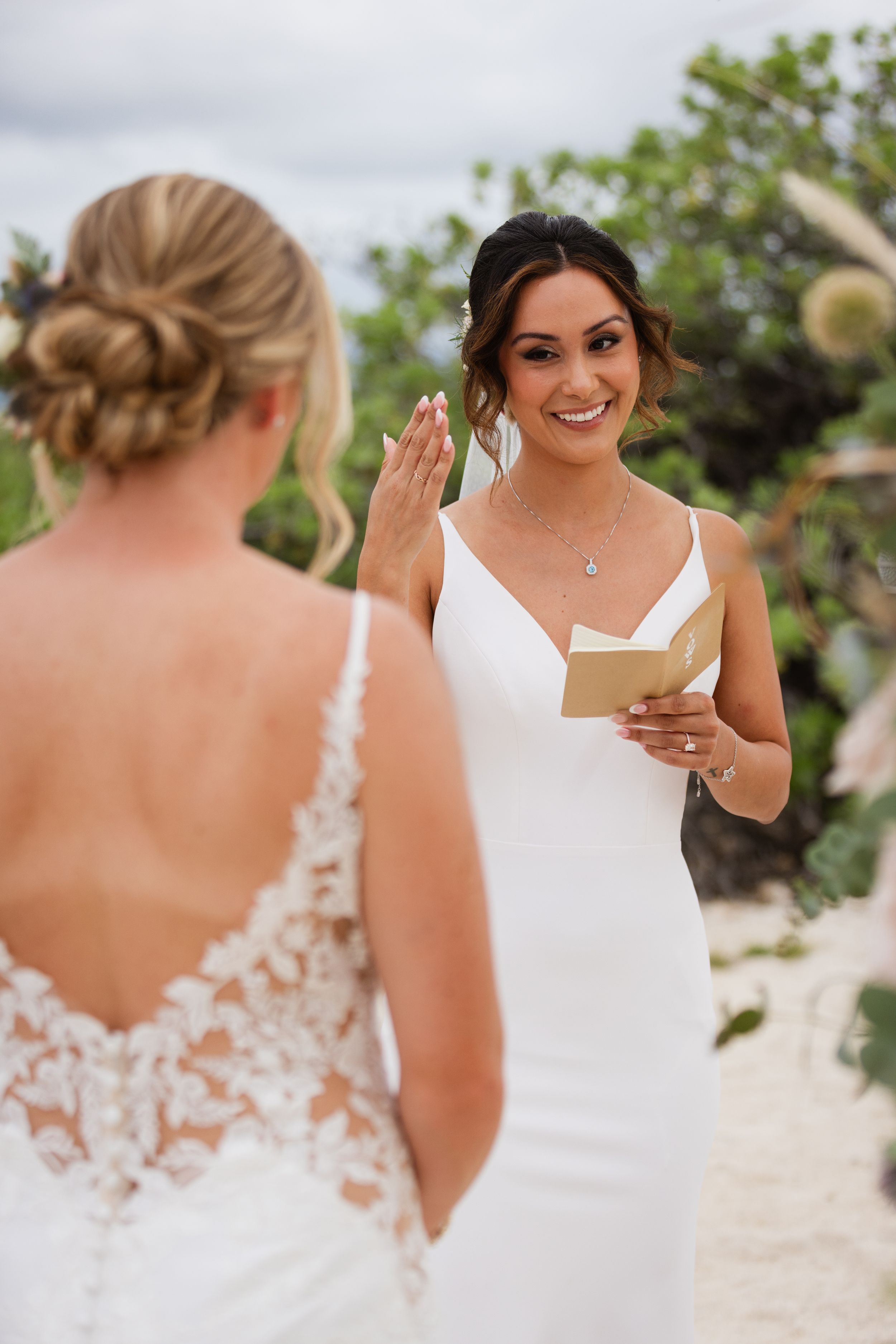 Smiling bride reads vows from a small book while facing her partner at an outdoor ceremony.