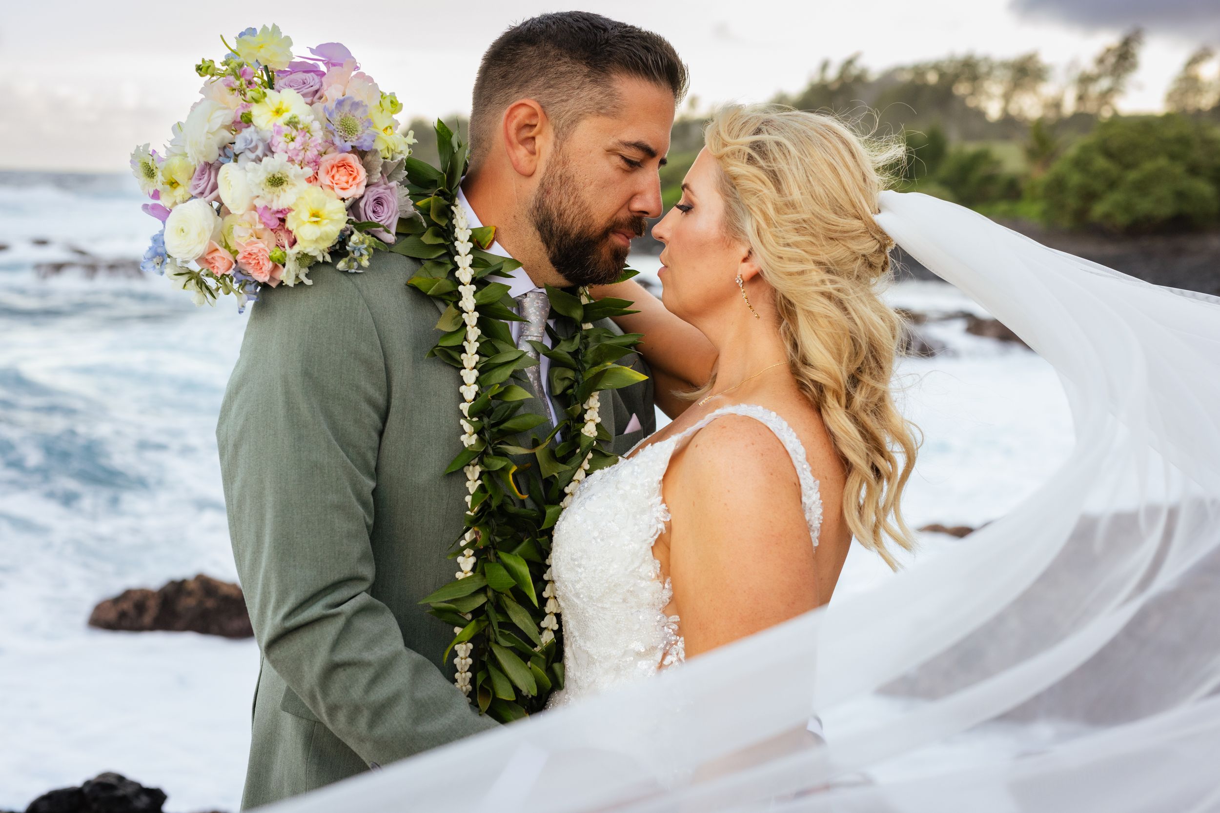 Close-up of couple nearly kissing with colorful bouquet and green lei on coastline.