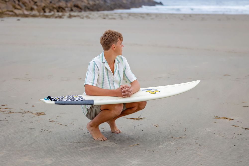 Surfer crouching on beach examining white surfboard with rocky coastline in background.