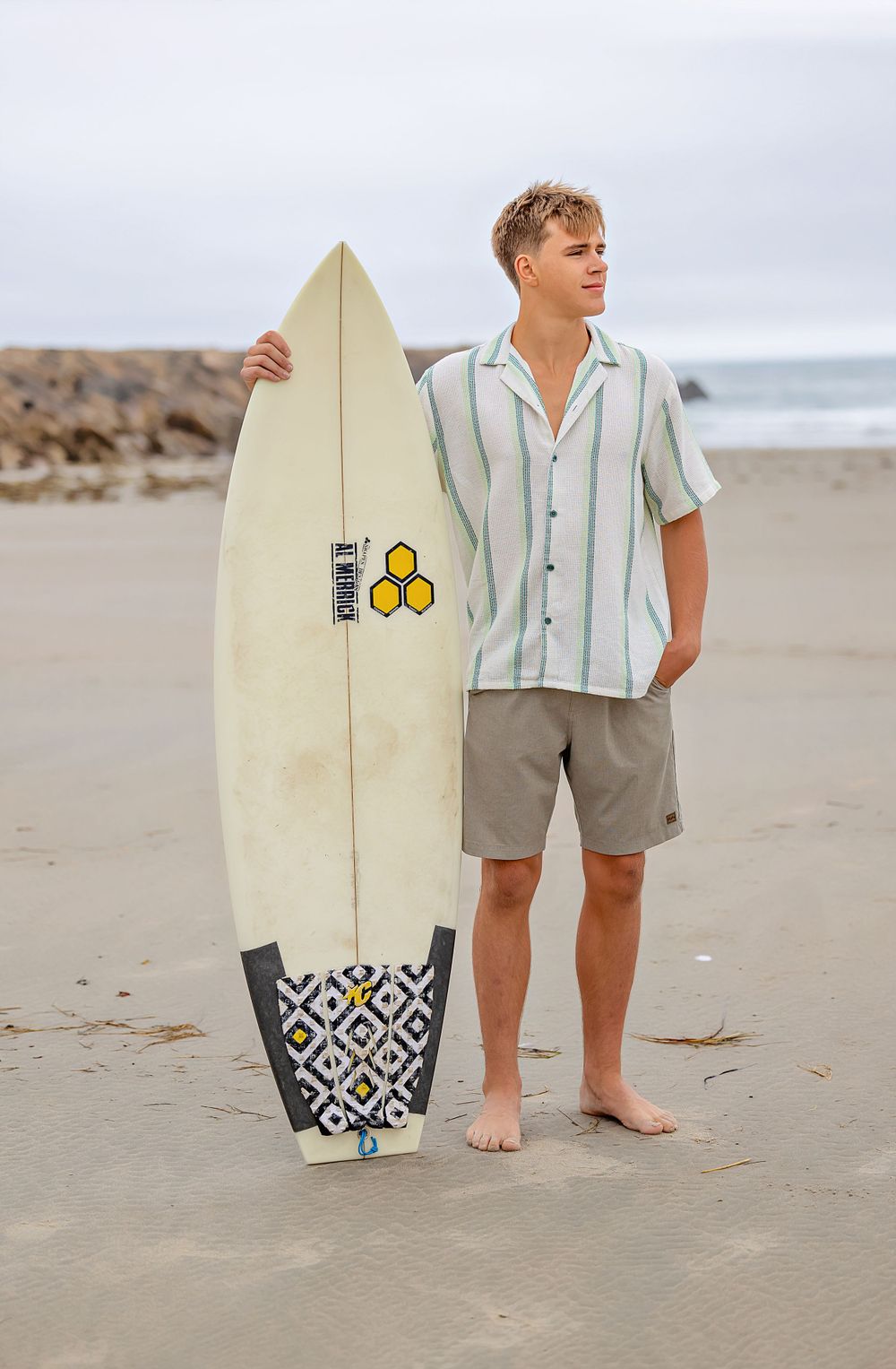 A casual beachside pose with a surfboard featuring striped shirt and khaki shorts.