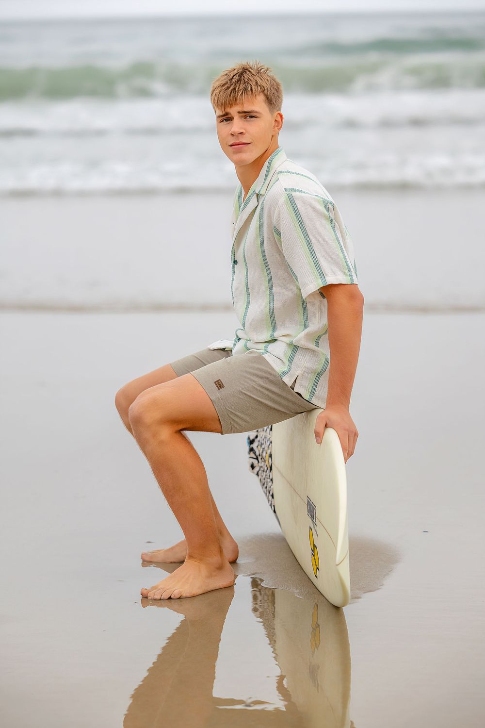 Casual beachwear style with striped button-down shirt and surfboard on sandy beach.