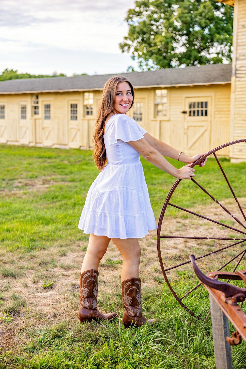 White summer dress and cowboy boots by an antique wagon wheel in a rustic setting.