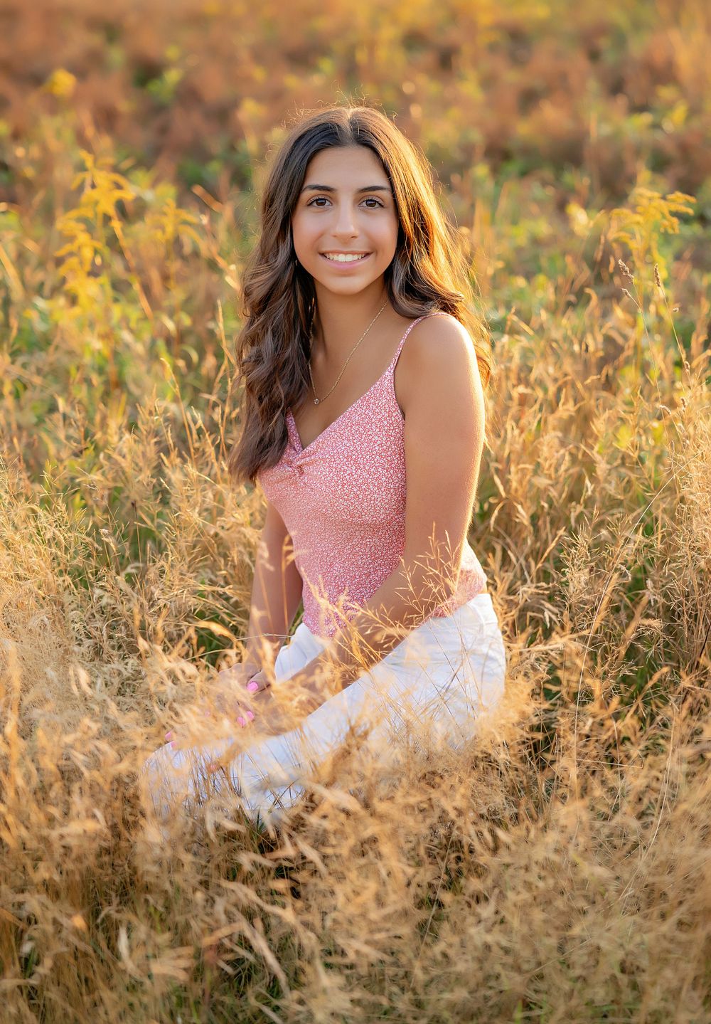 Portrait of a person in a pink top sitting in tall golden grass during sunset.