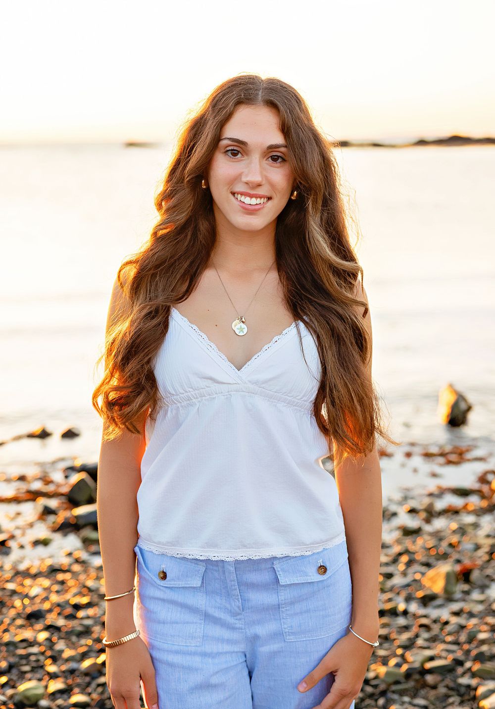 Woman in white sleeveless top and light blue shorts poses on rocky beach at sunset with ocean in background.