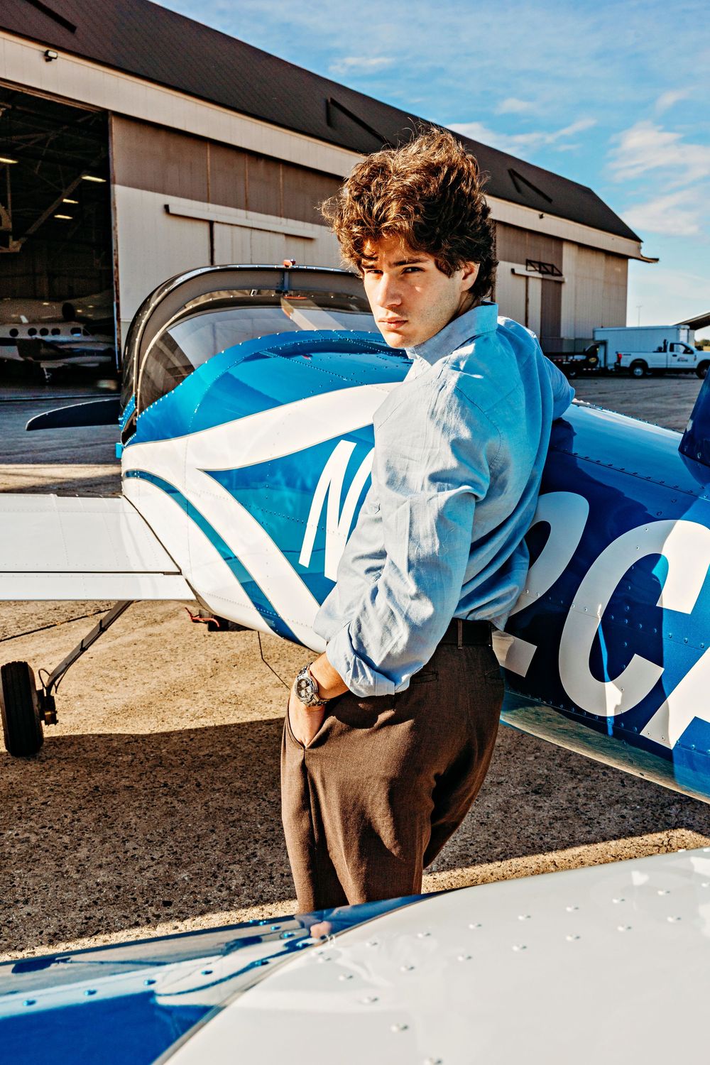 A young pilot stands next to a small aircraft on the tarmac wearing a white jacket and brown pants.