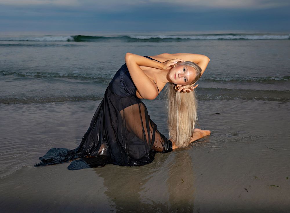 Artistic beach portrait in black flowing dress at sunset with waves in background.