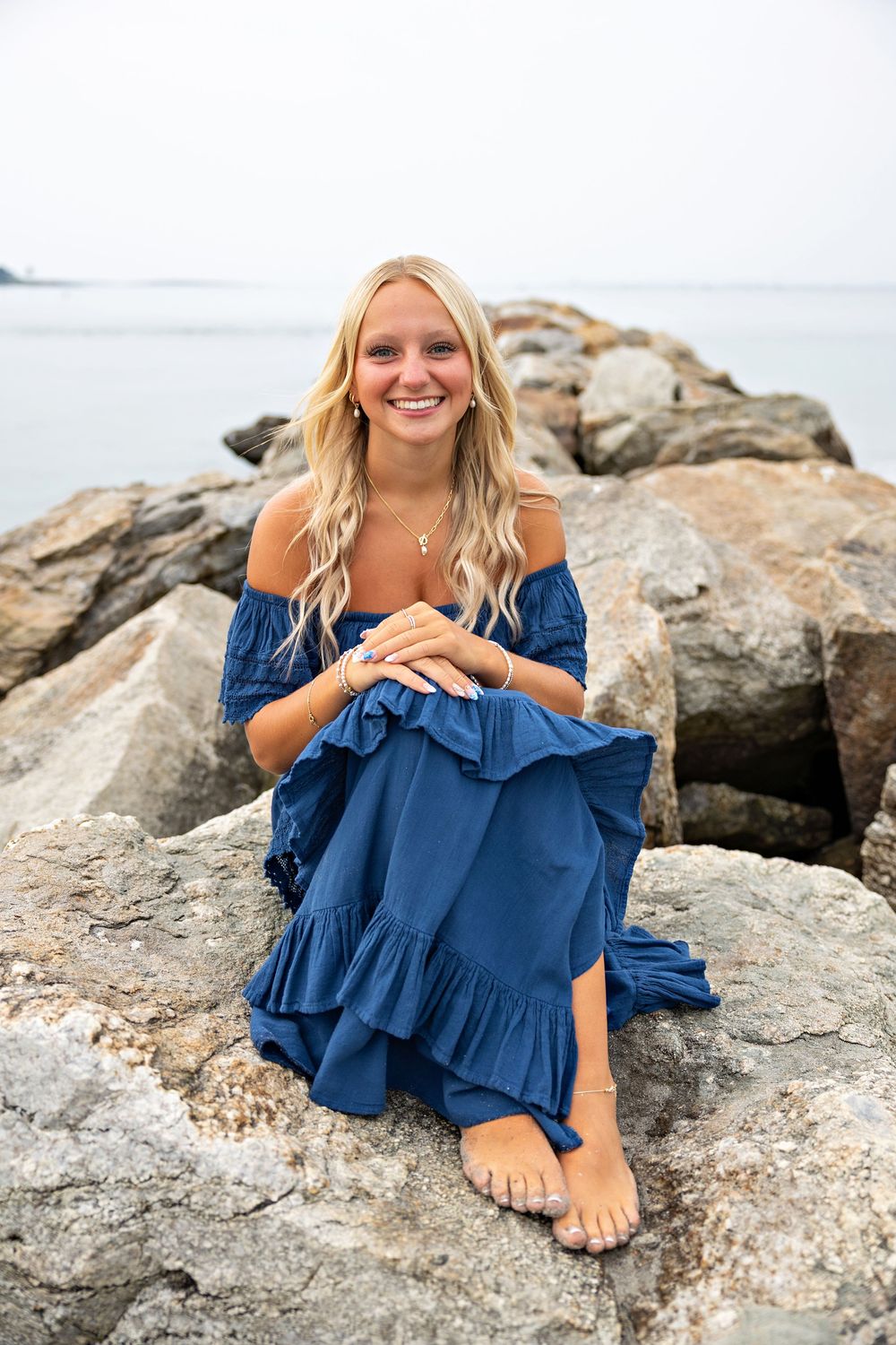 A person in a navy blue ruffled dress poses casually on coastal rocks near the ocean.
