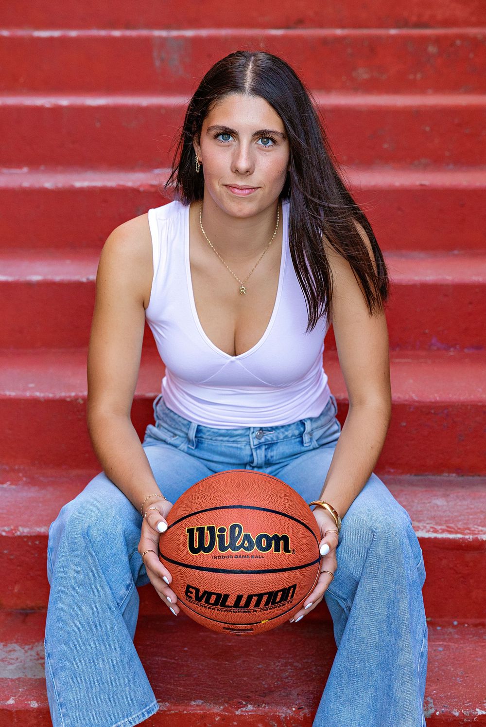 Basketball player in white tank top and jeans sits on red steps holding a Wilson Evolution basketball.