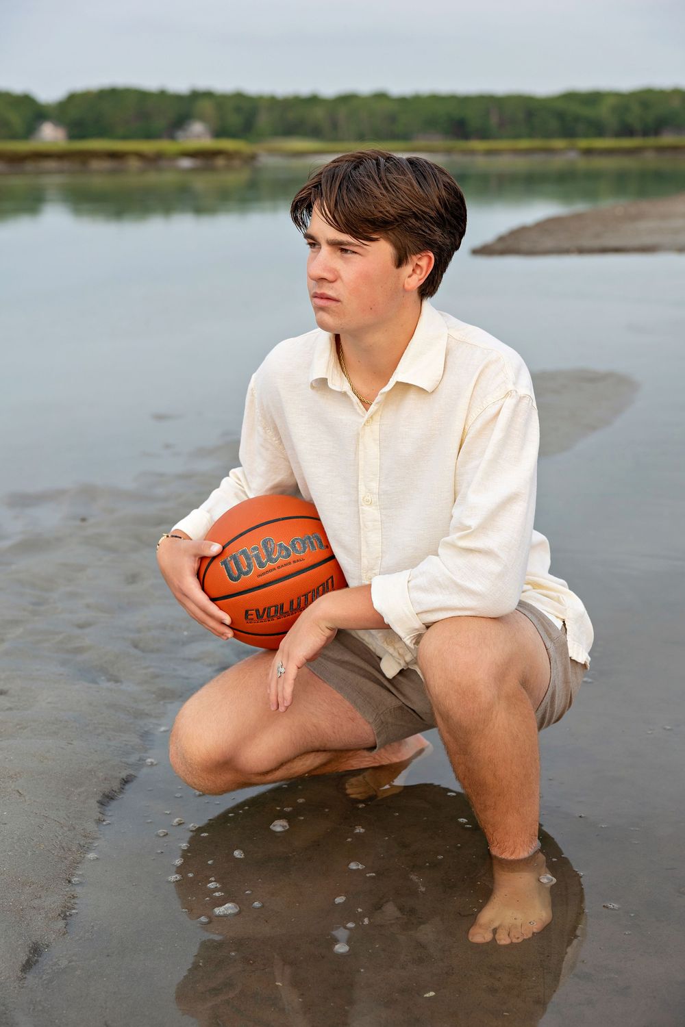 A person in a white shirt crouches on a beach holding a basketball while looking out over a lake.