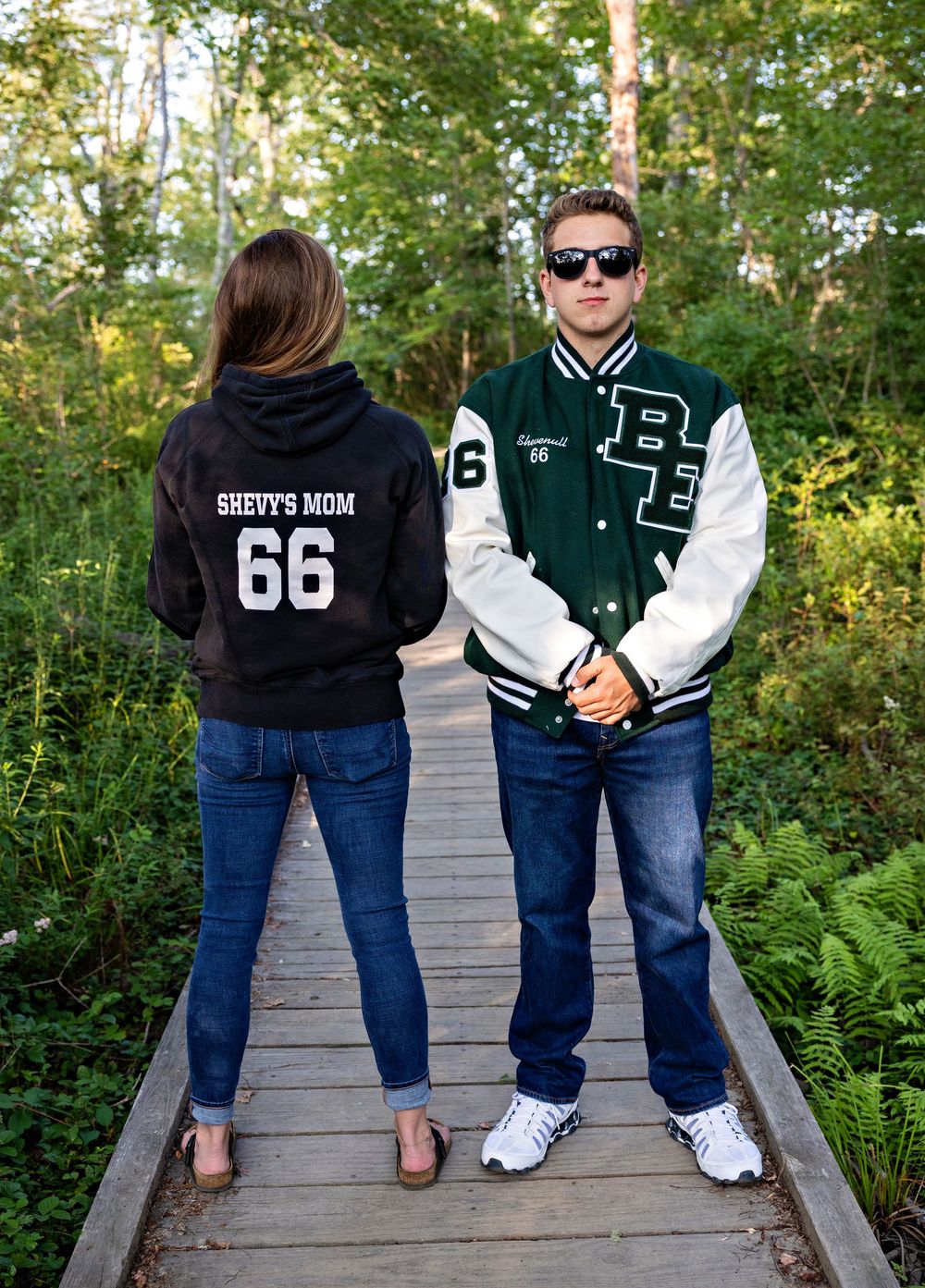 Two people wearing varsity jackets and jeans stand on a wooden boardwalk path surrounded by green foliage.