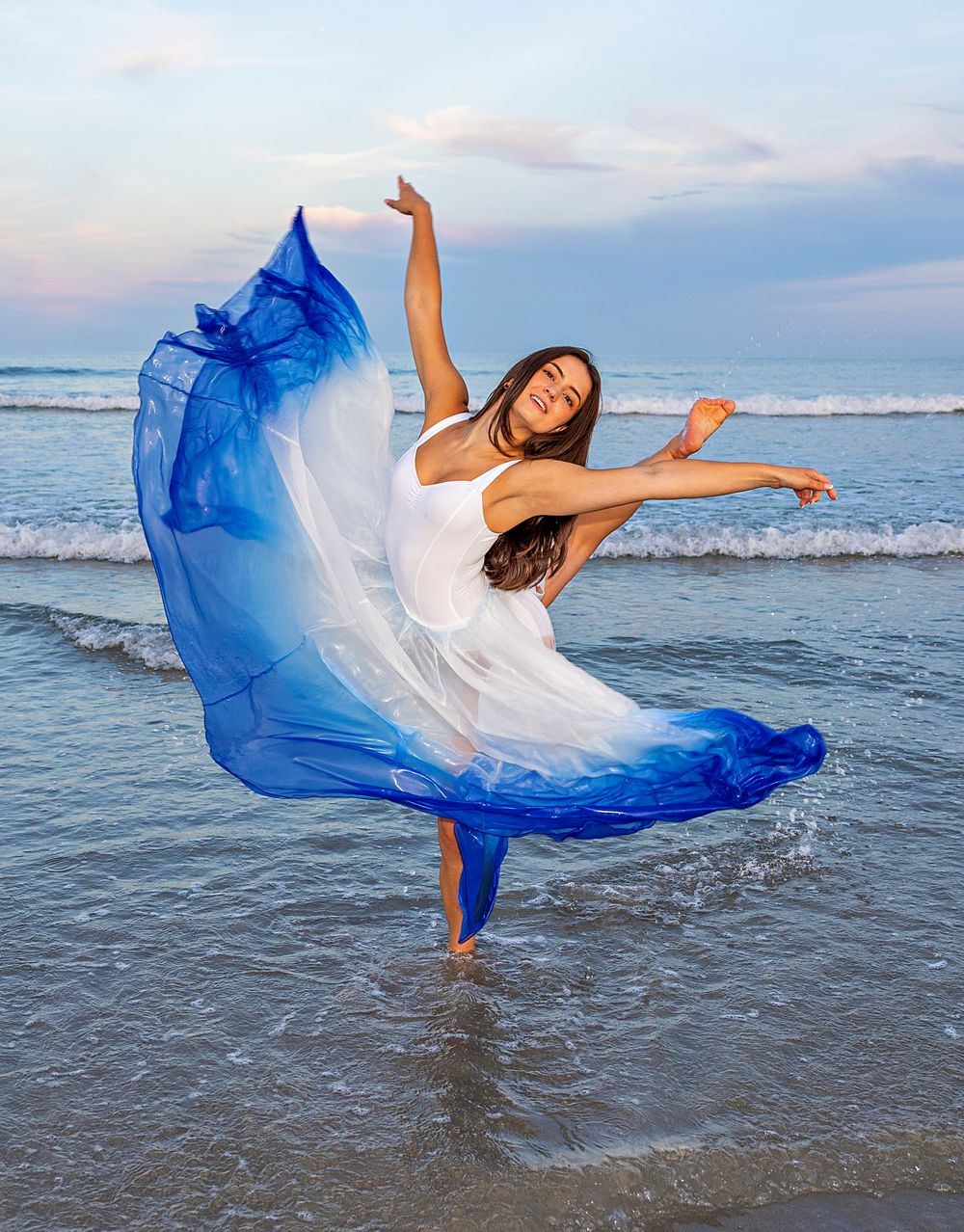 Dancer in flowing blue and white dress performs graceful movements at the ocean's edge during sunset.