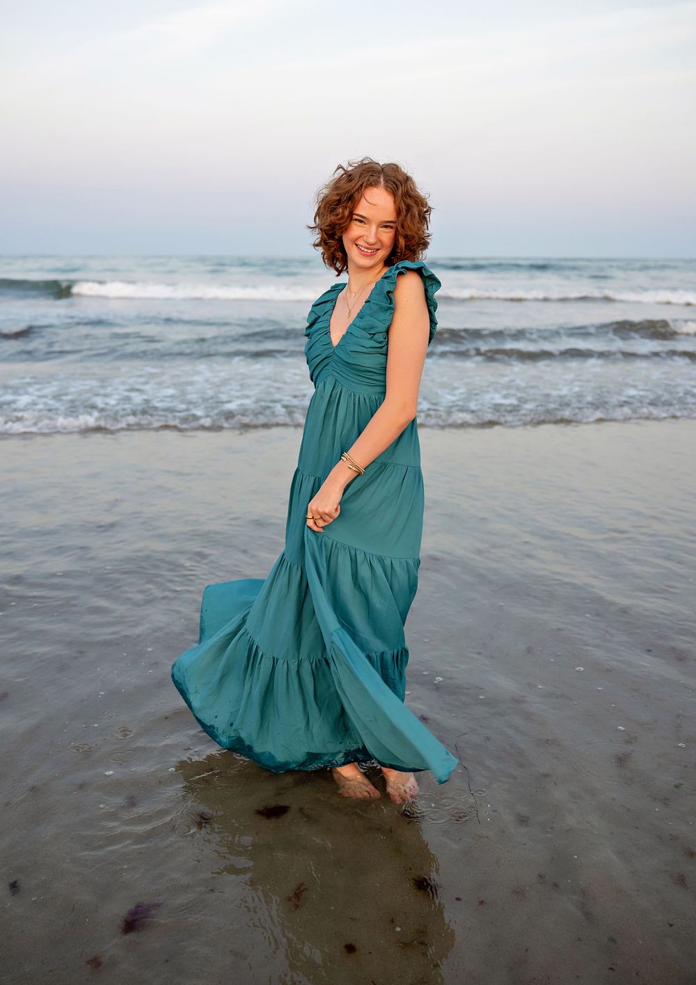 A figure in a flowing teal dress twirls and dances on a misty beach at sunset, creating a playful and carefree scene.