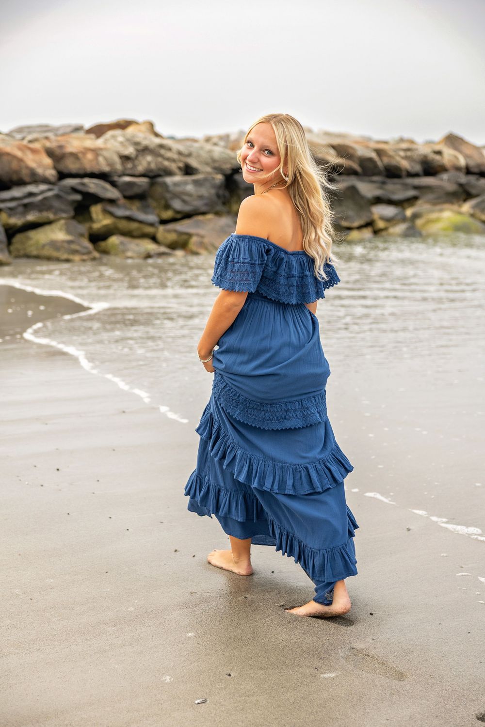 A woman in a flowing blue off-shoulder dress walks barefoot along a rocky beach shoreline.