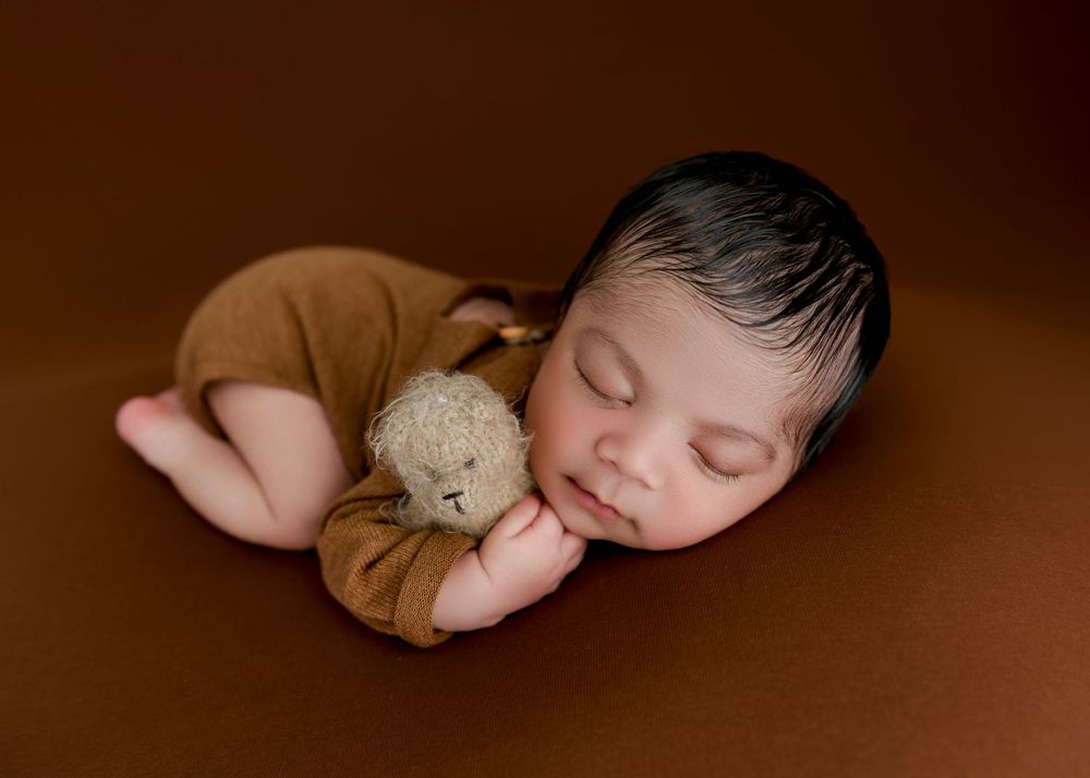 Sleeping newborn in brown outfit curled up with small teddy bear on dark brown backdrop during studio photography session.