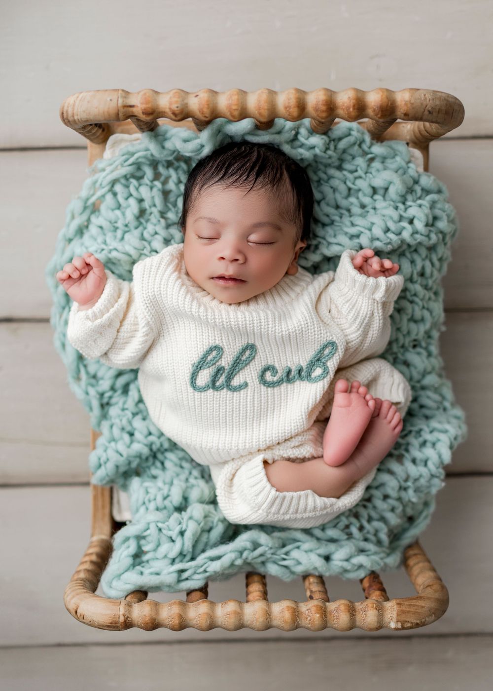 A sleeping infant wearing a white sweater lies on a chunky knit turquoise blanket in a wooden basket.