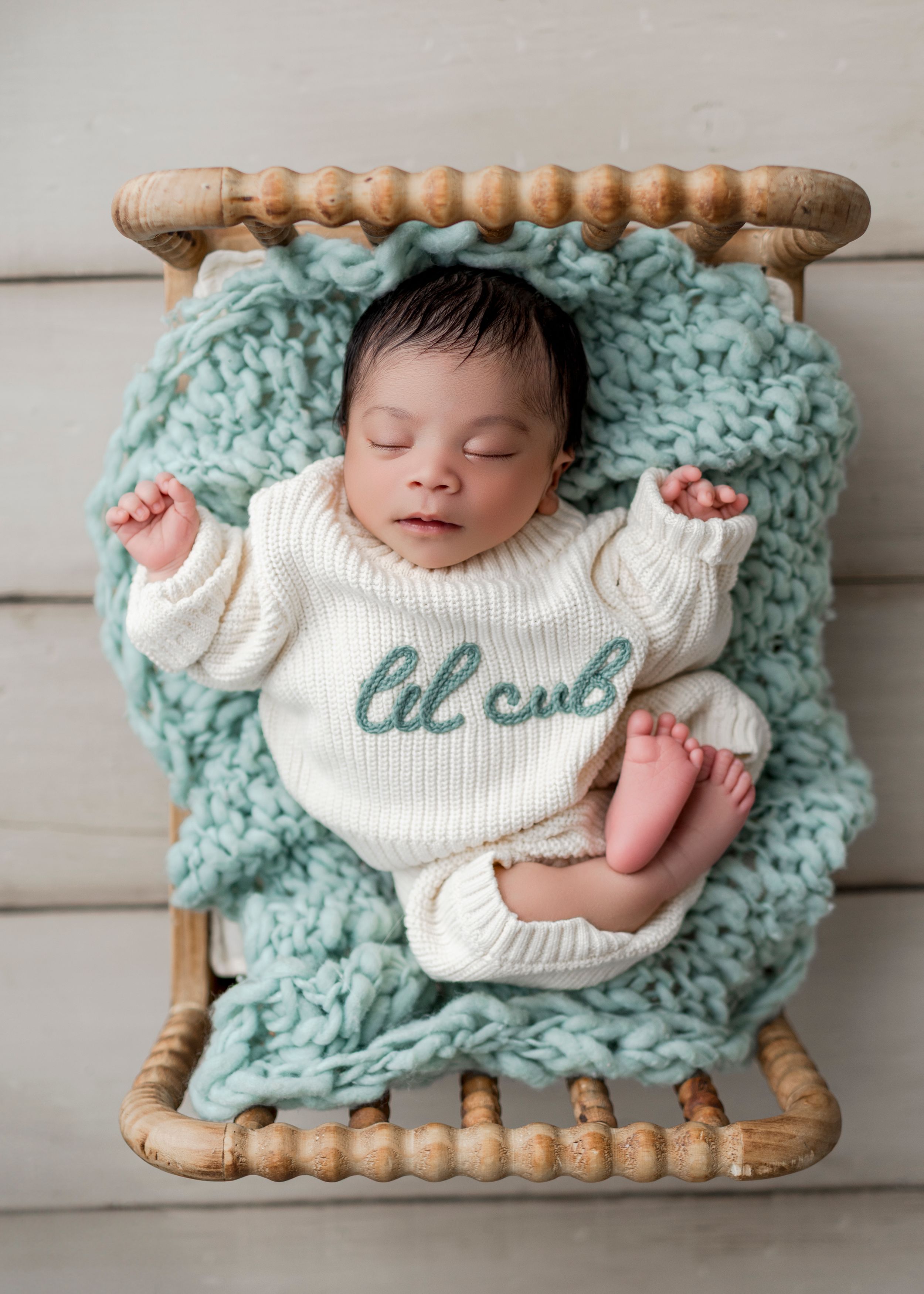 A sleeping infant wearing a white sweater lies on a chunky knit turquoise blanket in a wooden basket.