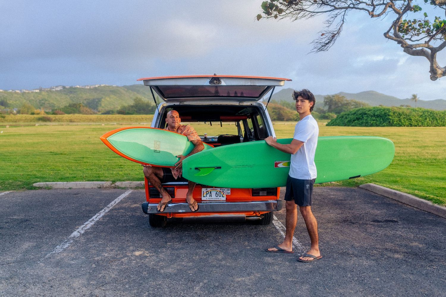 Two people loading colorful surfboards into vintage van at scenic Hawaii parking lot with mountains.