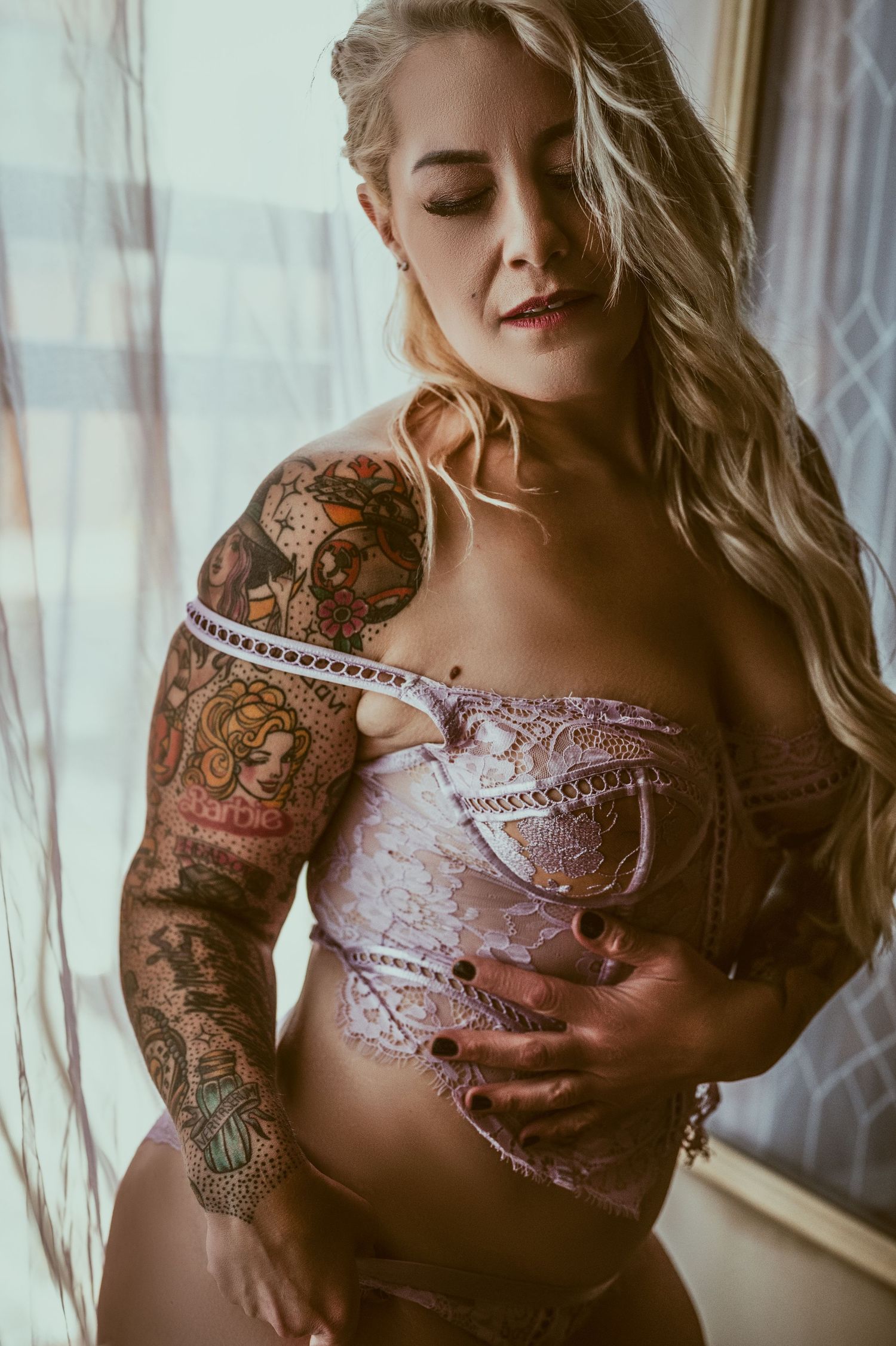 Artistic boudoir photo with intricate sleeve tattoos and delicate lace lingerie in natural window light.