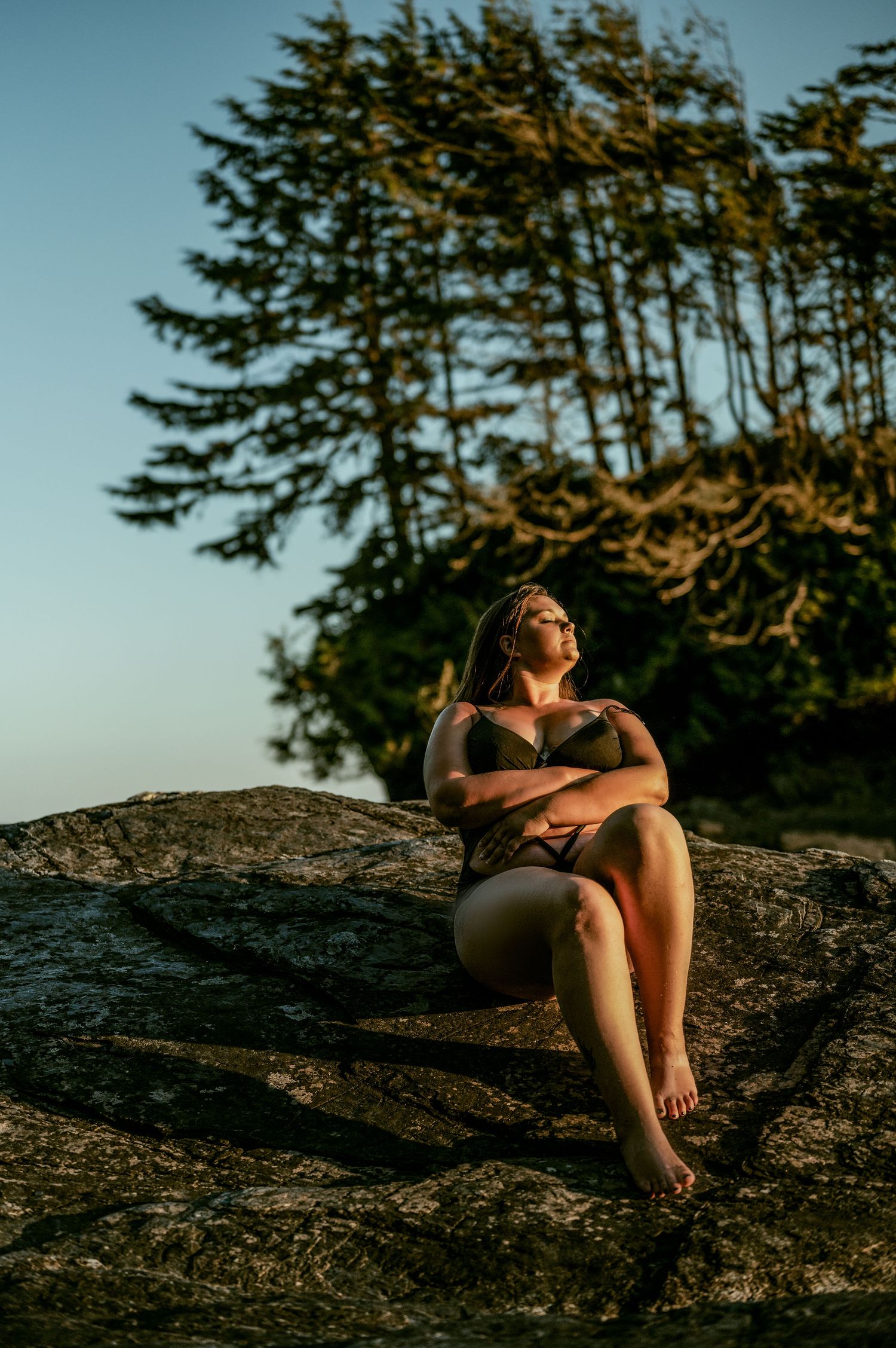 Figure sitting on a rocky outcrop at sunset with evergreen trees silhouetted against golden light in the background.