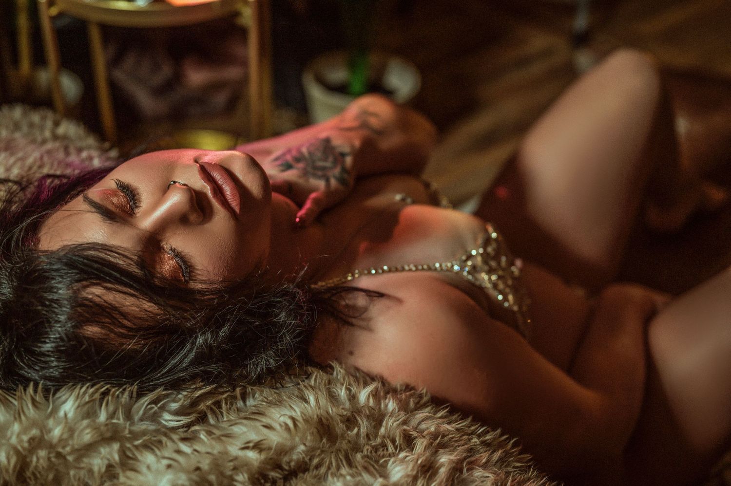 A person lies on plush fur fabric in moody red lighting, wearing glamorous jewelry and displaying visible tattoos.