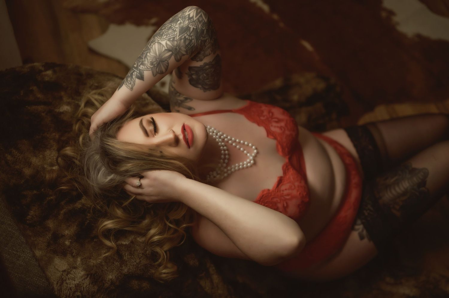 Artistic boudoir photograph with red lingerie and pearl accessories in warm lighting.