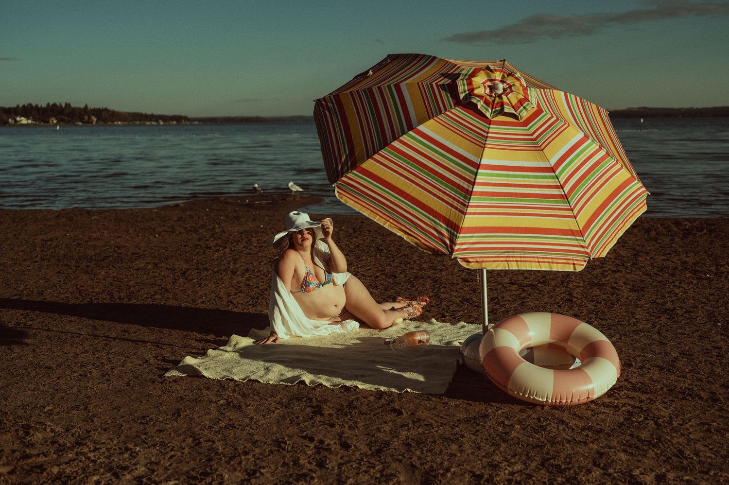 Woman relaxing on beach towel under striped umbrella with inflatable ring, photographing the calm ocean at sunset.