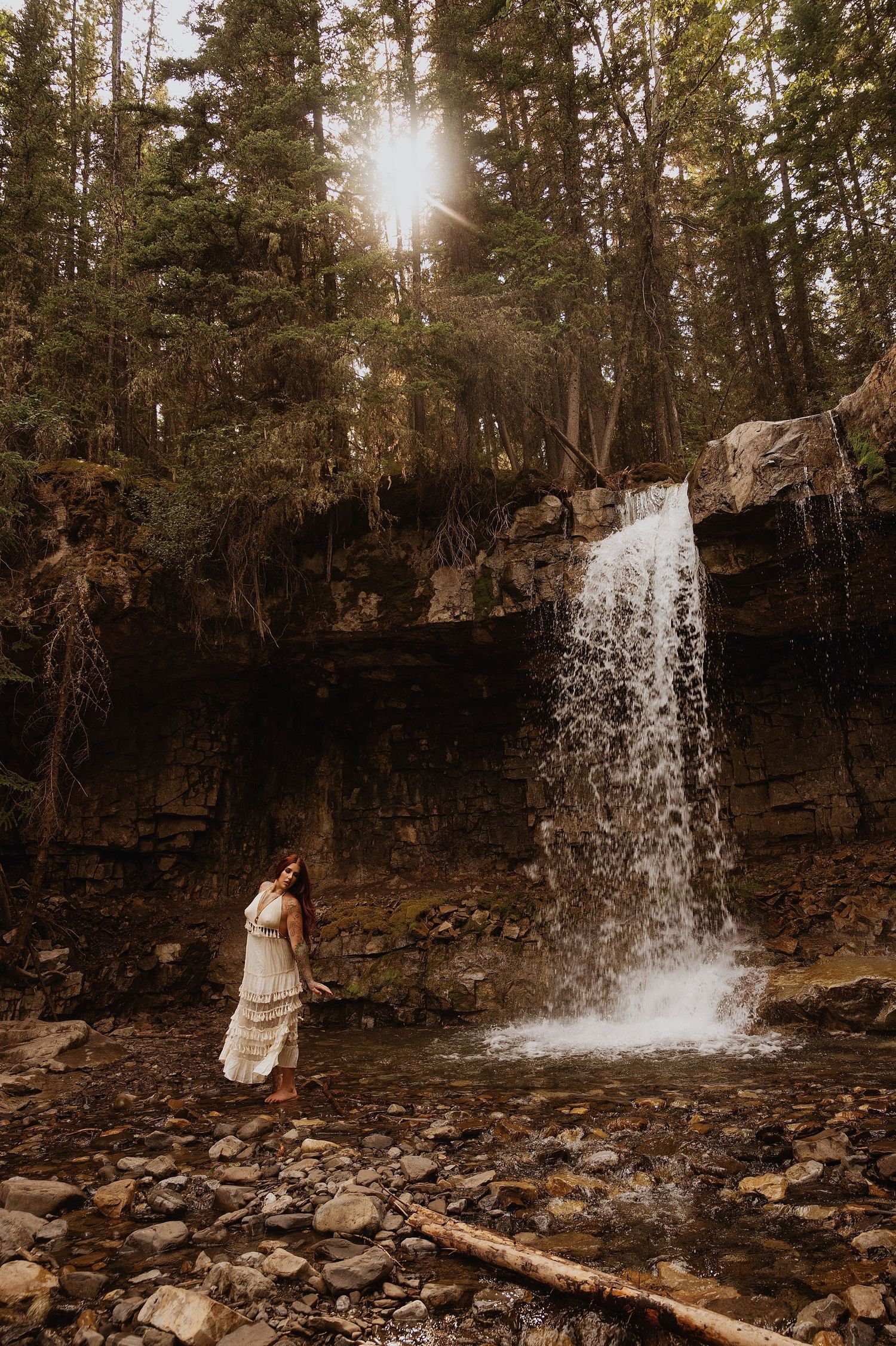 Woman in white dress standing beside a cascading waterfall in a lush forest with sunlight streaming through the trees.