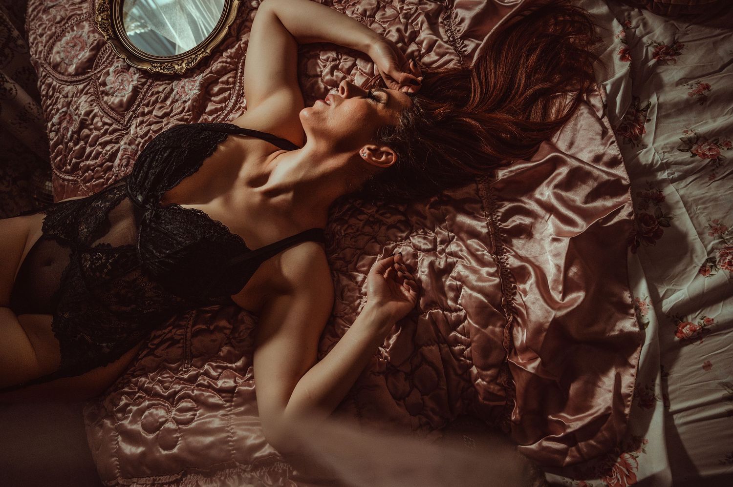 Woman in black lace lingerie lying on pink satin bedding in a moody, romantic boudoir photography setting.