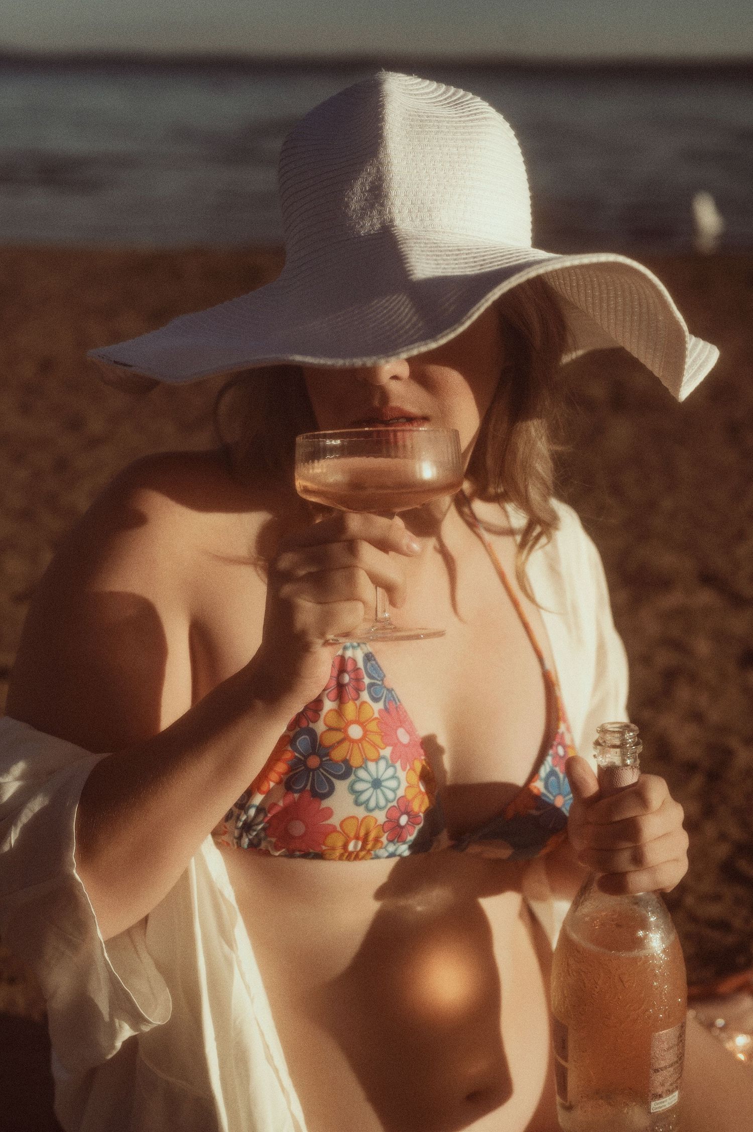 Woman in floral bikini and white sun hat sipping rosé wine on a sunny beach.