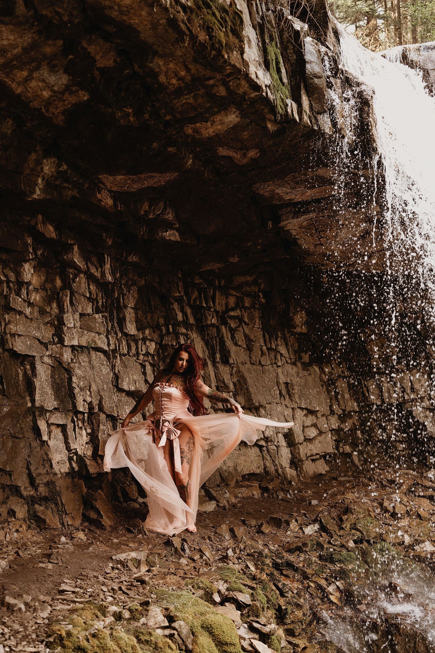 Two women in flowing gowns pose dramatically against a rocky cliff beside a cascading waterfall.