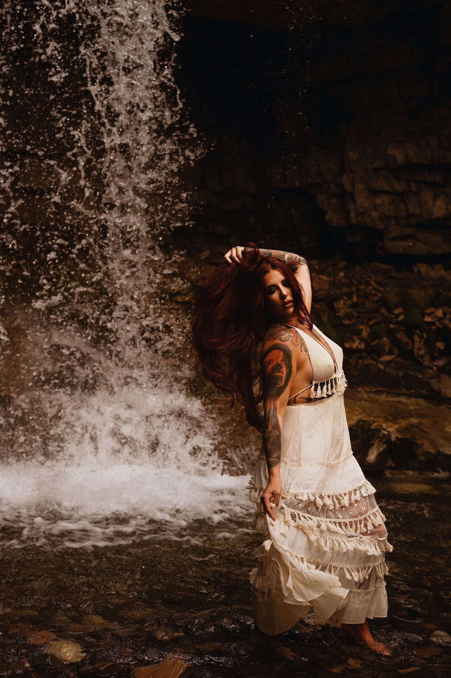 Woman in white ruffled dress poses gracefully before a cascading waterfall, her red hair flowing, showcasing a tattoo.