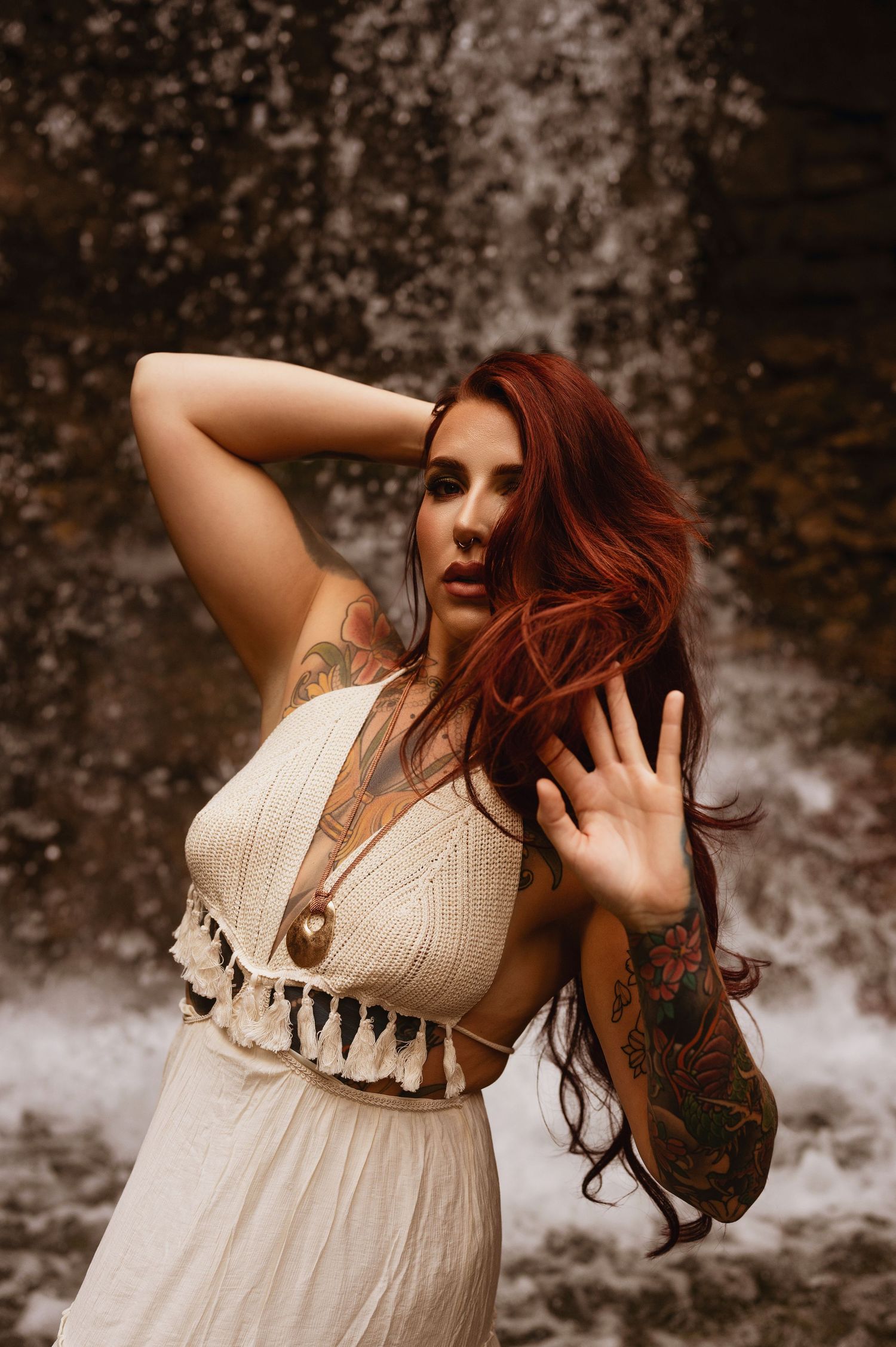 Red-haired tattooed woman in white boho dress posing gracefully in front of a waterfall.