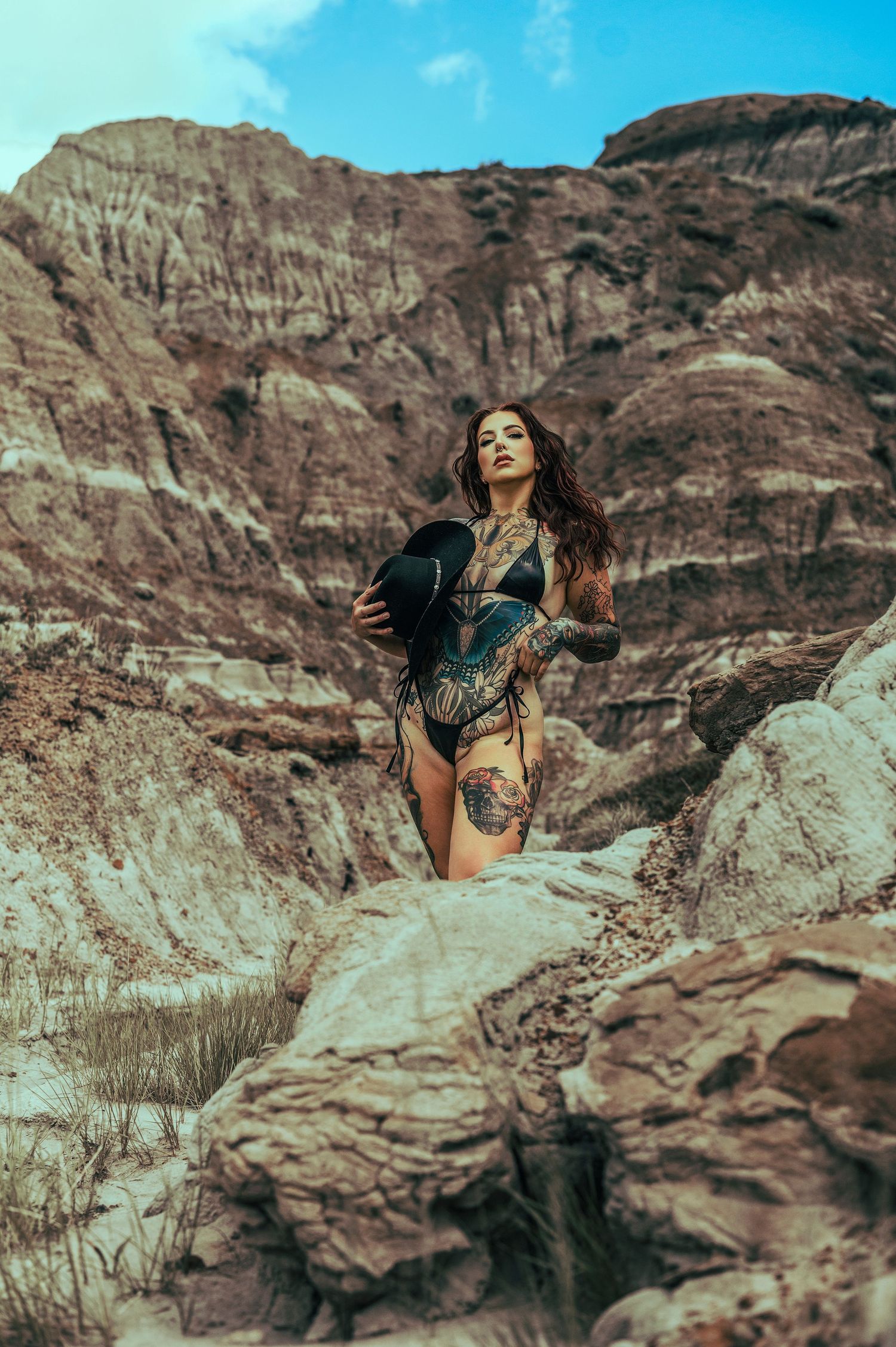 Heavily tattooed woman in black swimsuit posing on rocky desert canyon formations under a blue sky.