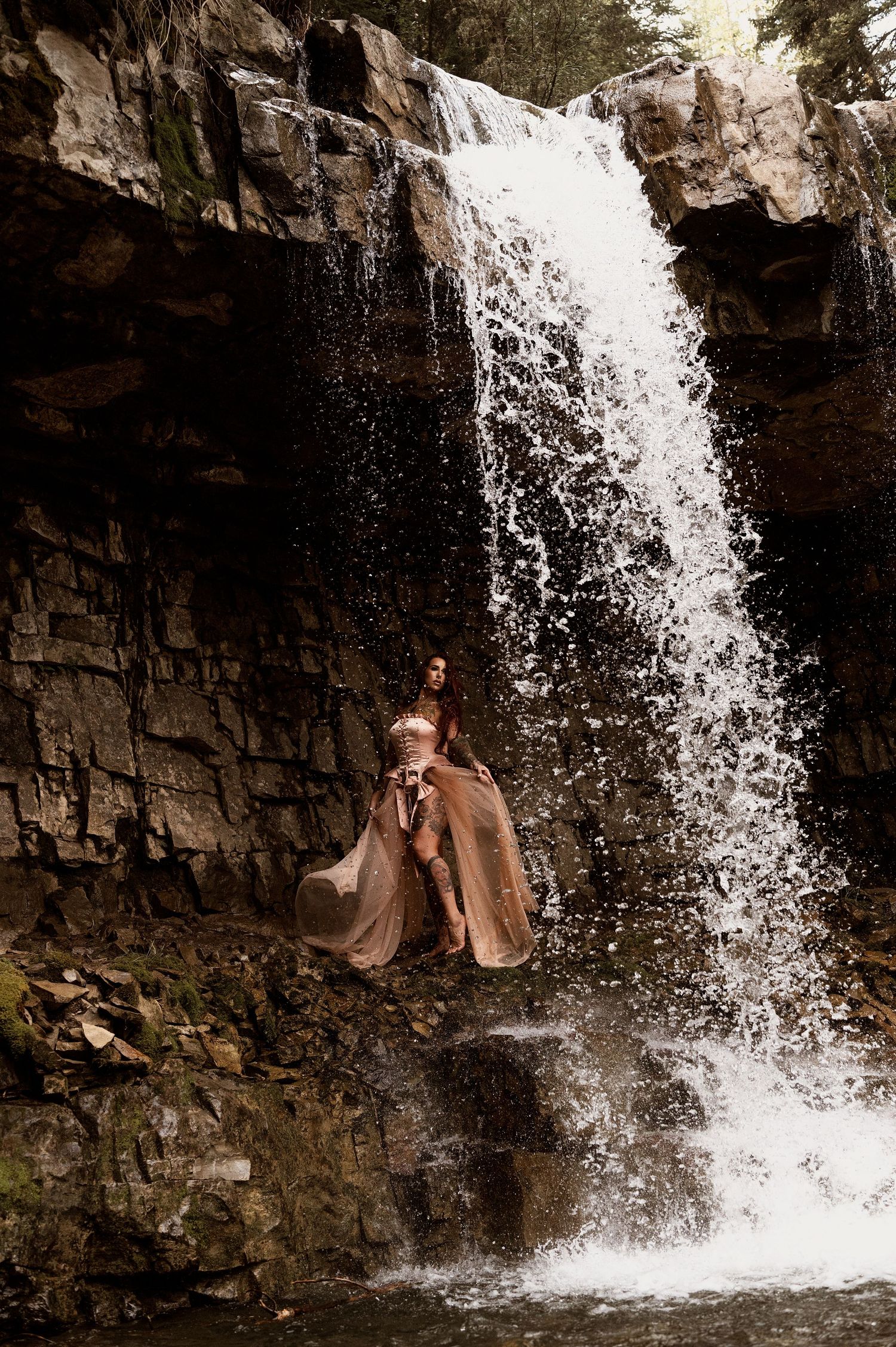 Woman in flowing rose gold gown standing before a cascading waterfall on rocky terrain.