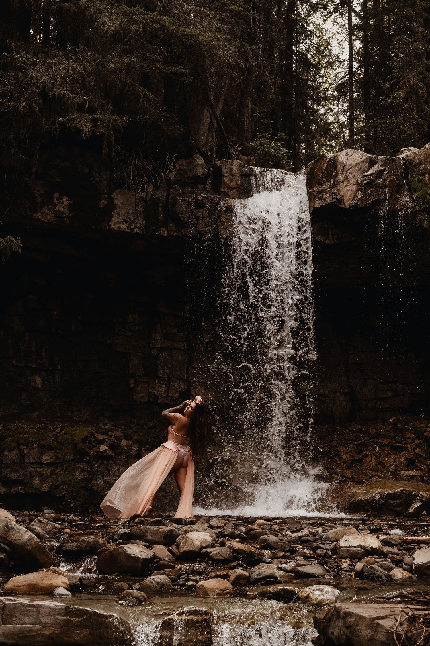 Woman in flowing pink dress poses gracefully before a cascading waterfall surrounded by rocks and evergreen trees.