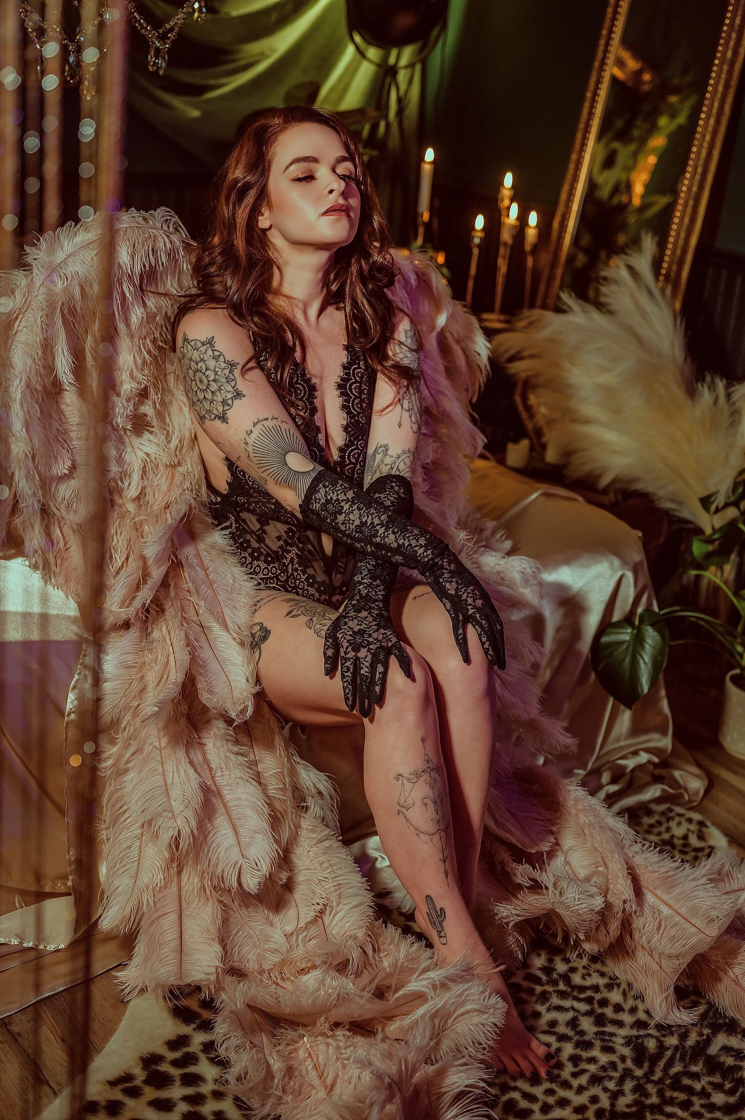 Tattooed woman in black lace bodysuit and fur coat poses elegantly by candlelight in a glamorous boudoir setting.