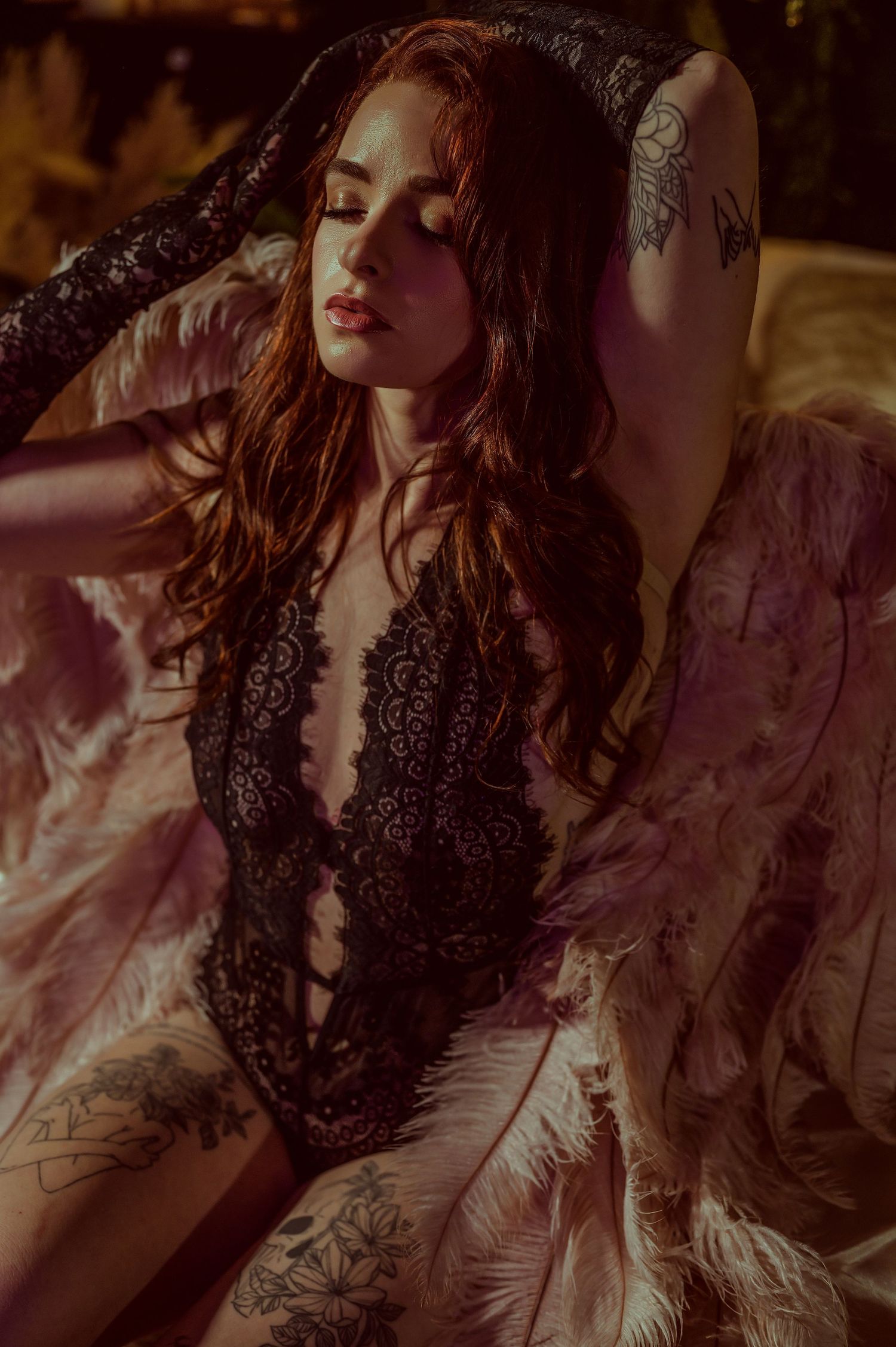 A tattooed redhead woman poses in black lace lingerie surrounded by feathers in a moody, dramatic boudoir setting.