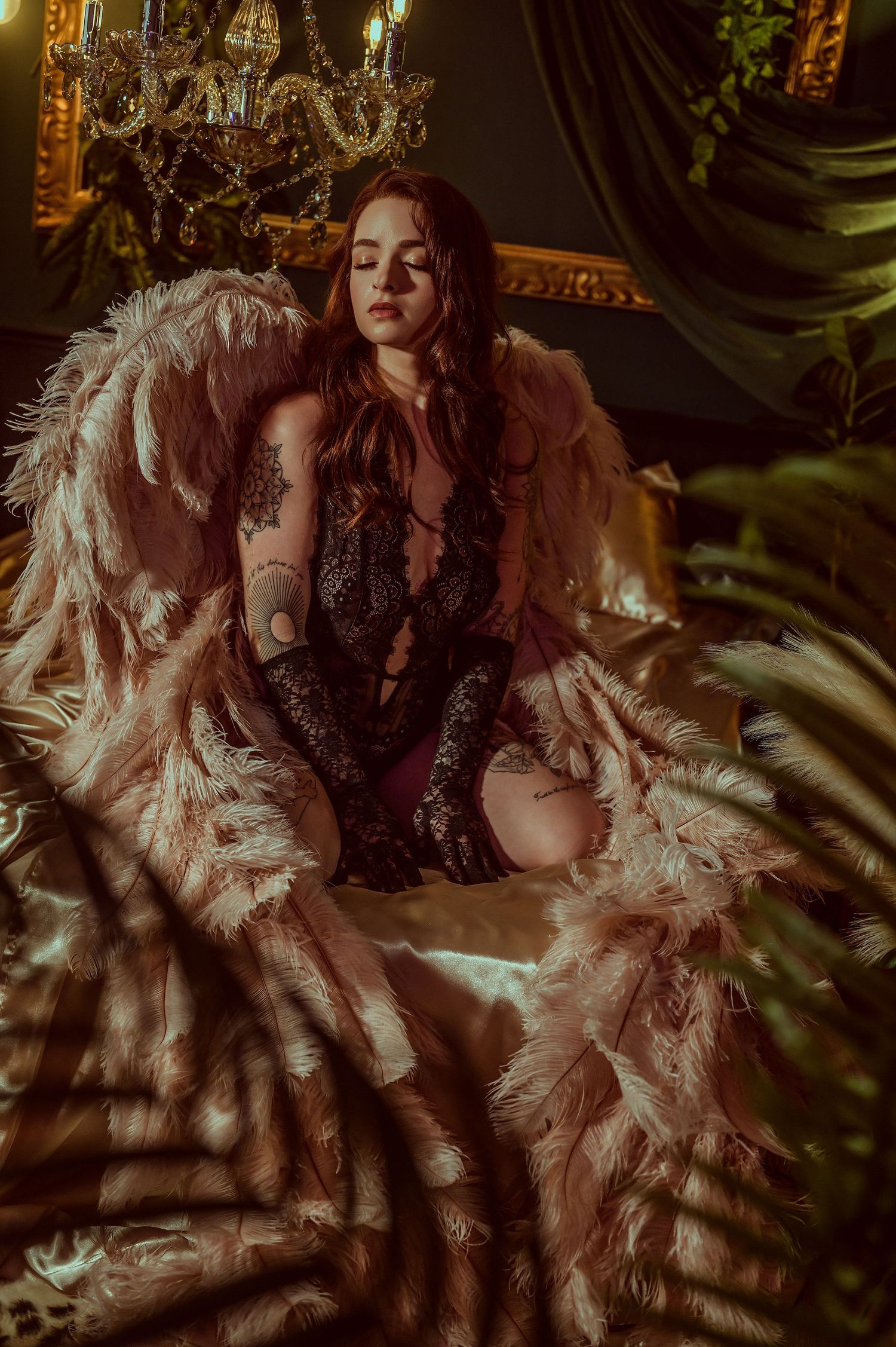 A tattooed woman in black lace lingerie sits on a fur chair surrounded by lush greenery and a golden chandelier.
