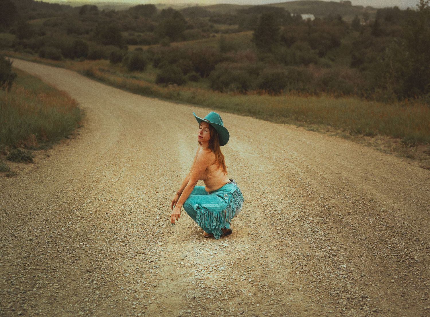 A woman in a teal hat and fringed skirt crouches on a rural dirt road surrounded by green hills.