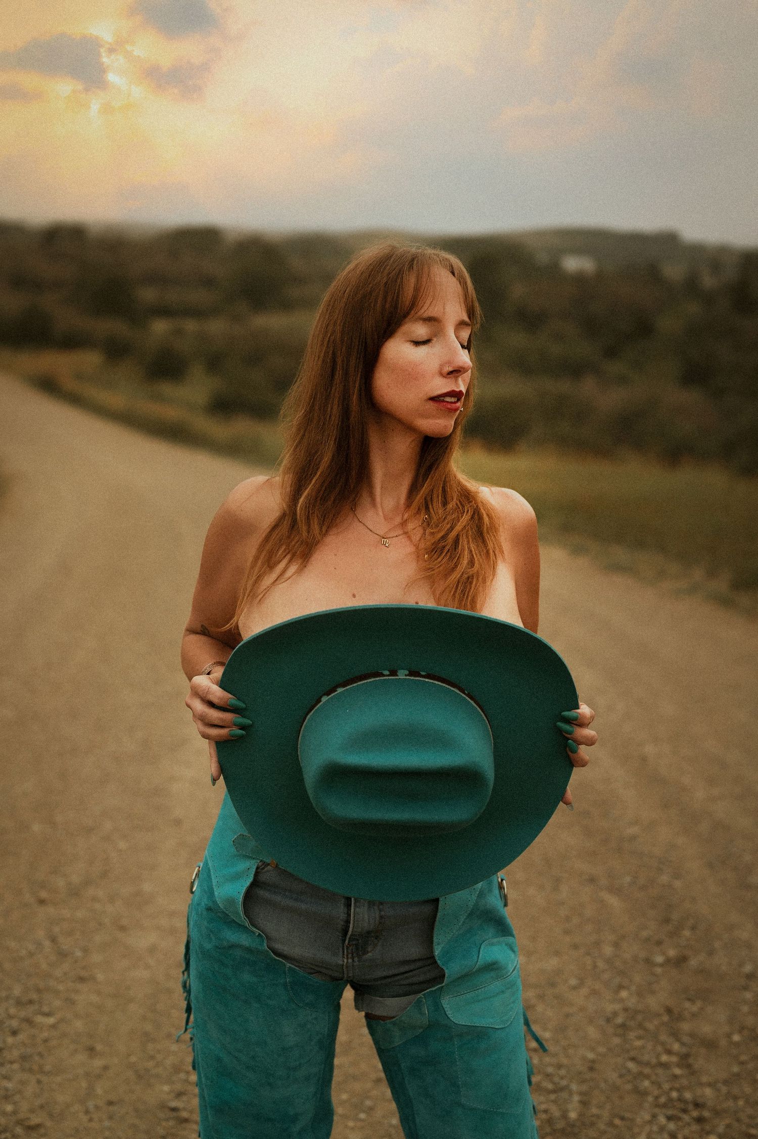 Woman holding teal cowboy hat on rural road at sunset wearing turquoise chaps.